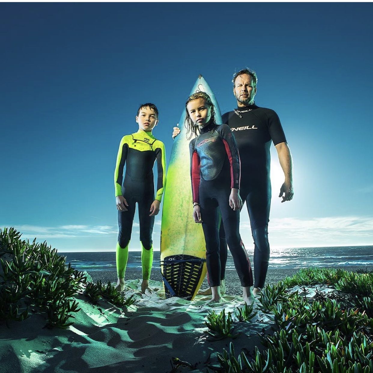 William Sanford in a wetsuit on the beach at sunset with his two kids and their surfboards