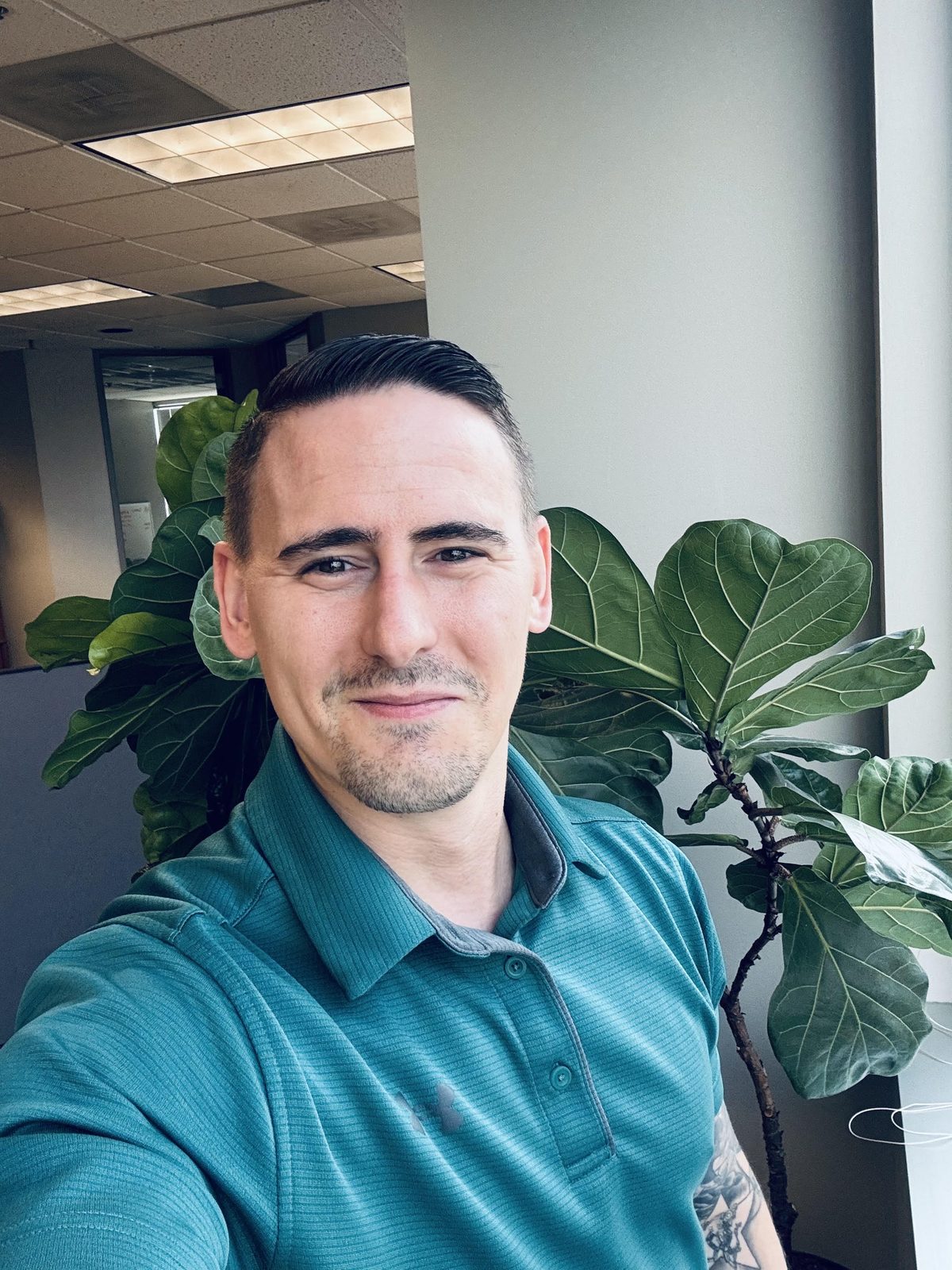 Zach Nuss, owner of Nuss Fitness and Strong Will Press, smiling in a teal polo shirt in his Kansas City office